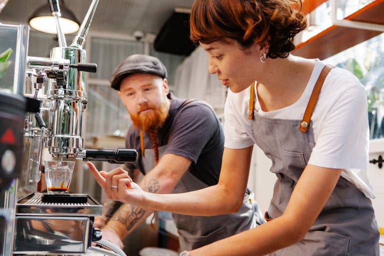 Crop female cafe employee speaking with tattooed male coworker against modern espresso maker pouring coffee into glass